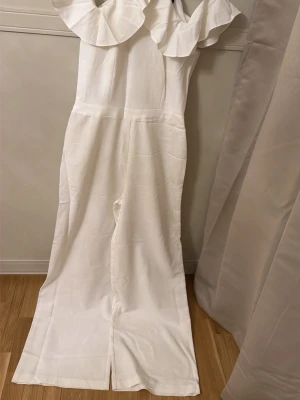 Vit jumpsuit med volangkrage - Snygg vit jumpsuit med breda ben och stor volangdetalj runt halsen. Ärmlös modell med hög hals och dragkedja i ryggen. Perfekt för dig som vill sticka ut med en stilren och trendig look. Materialet känns mjukt och har ett fint fall.. Använts endast en gång.