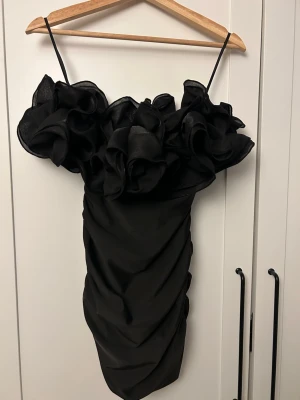 Svart kort klänning - Svart kort tubklänning med stora volangdetaljer upptill. Klänningen har en figurnära passform och passar bra till festliga sammanhang. Materialet är stretchigt och har en dragkedja på sidan