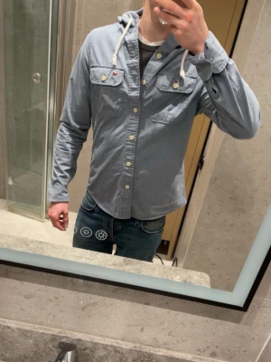 Hollister overshirt  - Snygg ljusblå overshirt från Hollister med huva och vita dragsnören. Två bröstfickor med knappar och klassisk knappstängning framtill. Mjuk bomullskänsla och avslappnad passform, perfekt för lager på lager. Liten Hollister-logga på bröstet.