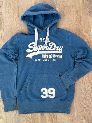 Blå hoodie från Superdry med tryck - Snygg blå hoodie från Superdry med vit logga och japansk text på bröstet samt siffran 39 på fickan. Hoodien har huva med vita dragsnören och en stor magficka. Tillverkad i mjukt bomullsmaterial, perfekt för en avslappnad streetwear-look. Två fläckar finns+lite på loggan, en fläck syns knappt så fråga om bild om du vill ha det. Märkt som XS men skulle säga att den är en S.