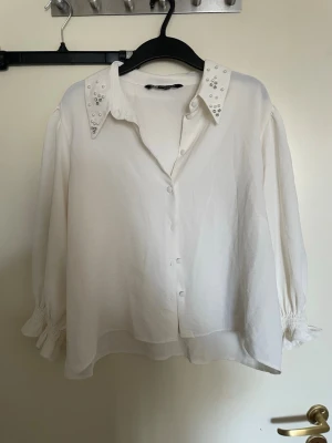 Vit skjorta med pärldetaljerad krage - Stilren vit skjorta med pärldetaljer på kragen och knappar framtill. Skjortan har långärmade puffärmar med smock vid ärmsluten och en lös passform som ger en avslappnad men chic look. Perfekt för dig som gillar detaljer och vill ha något extra till outfiten.