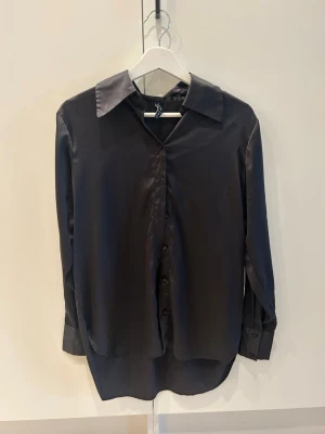 Svart silkesliknande skjorta - Stilren svart skjorta med lång ärm och klassisk krage. Skjortan har en slät, glansig yta som ger en lyxig känsla och stängs med knappar framtill.