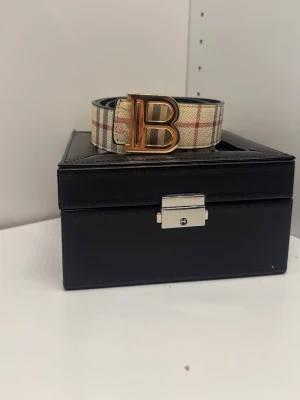 Rutmönstrat bälte med guldspänne - Snyggt bälte från Burberry med klassiskt rutmönster i beige, rött och svart. Bältet har ett stort guldfärgat B-spänne som ger en lyxig känsla. Perfekt accessoar för att lyfta vilken outfit som helst.