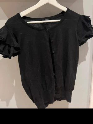 Svart kofta med volangärmar - Kortärmad svart kofta med volangärmar och rund halsringning. Knäpps framtill med små knappar och har ribbad nederkant. Perfekt för dig som gillar en söt och trendig stil. Materialet känns mjukt och lätt.