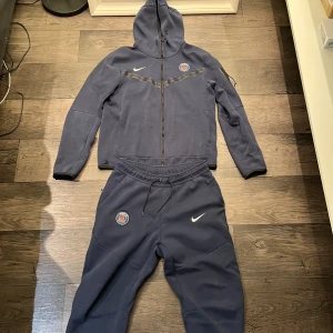 PSG nike tech - Säljer en mörkblå tech fleece från Nike x PSG med hel dragkedja, huva och klubbmärke på bröstet. Tröjan har långa ärmar, snygg detalj med svart band över bröstet och en ficka med dragkedja på ärmen.Tröjan har ett litet hål på höger sidan vid höften, men det märker man knappt av. Perfekt för dig som gillar fotboll och streetwear.