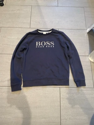 Mörkblå sweatshirt från Hugo Boss - Cool mörkblå sweatshirt från Hugo Boss med BOSS-logga i vitt på bröstet. Tröjan har rund halsringning, långa ärmar och ribbade muddar. Perfekt för dig som gillar stilrena och klassiska plagg med en touch av premiumkänsla.