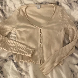 Beige ribbad kofta med pärlknappar - Jättesöt ribbad beige kofta med pärlknappar framtill och små volanger vid armslutet och nedre delen av koftan. Storlek L men liten i storleken så hade passat S/M.