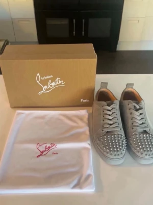 Grå Christian Louboutin sneakers med nitar - Grå Christian Louboutin med coola silverfärgade nitar på tån och klassisk röd sula. Skorna är gjorda i mocka och har en låg profil med vita skosnören. Perfekt för dig som vill sticka ut med exklusiva detaljer.