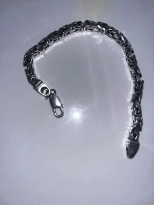 Kedjearmband i silverfärg - Snyggt armband i silverfärgad metall med en robust kedjedesign. Armbandet har ett klassiskt karbinlås och länkar med en flätad look som ger en cool och stilren känsla. Perfekt accessoar för att lyfta din outfit.