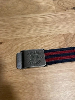 Randigt bälte från Hampton Republic - Snyggt bälte från Hampton Republic med metallspänne och ingraverad logga. Bältet är randigt i marinblått och rött, och är gjort i slitstarkt tygmaterial. Perfekt accessoar för att lyfta din outfit.