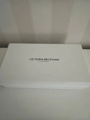 Vit box Victoria Beckham Eyewear - Säljer en vit rektangulär box från Victoria Beckham Eyewear. Boxen har en stilren design med logotyp på locket och är tillverkad i hård kartong med textilstruktur. Perfekt för förvaring av glasögon eller accessoarer.Mått 21 *12 cm, höjd 5 cm.