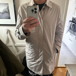 Schoffa Beige trenchcoat med svarta knappar - Stilren beige trenchcoat med klassisk krage och svarta knappar framtill. Långa ärmar med snygga knappdetaljer vid ärmslut. Rak passform och lätt material som ger en clean look. Perfekt för dig som gillar minimalistisk stil.