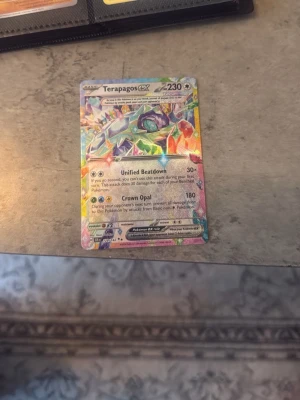 Terapagos GX Pokémonkort - Säljer ett färgglatt Terapagos GX Pokémonkort med holografisk yta. Kortet har 230 HP och attacker som Unified Beatdown och Crown Opal. Perfekt för samlare eller Pokémonfans som vill utöka sin kortlek. Kan diskutera pris