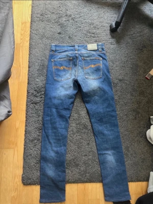 Blå slim fit jeans från Nudie Jeans - Säljer ett par snygga nudie jeans i färgen bay blue, byxorna är i gott skick och storleken 28/32.  Midje mått 38cm  Total längd 103cm  Benöppningen 16cm