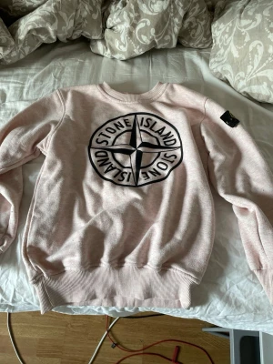 Stone Island rosa sweatshirt - Rosa sweatshirt från Stone Island med stort svart och vitt logotryck på bröstet och ikonisk patch på vänster ärm. Tröjan har rund hals, ribbade muddar och är tillverkad i mjukt material som passar perfekt för en avslappnad look. Storlek 164, billigare pris vid direkt köp 