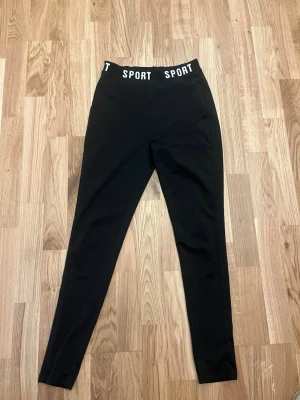 Svarta leggings med SPORT-resår - Snygga svarta leggings med hög midja och bred resår där det står 'SPORT' i vitt. Tight passform och stretchigt material som ger en sportig vibe. Perfekta till både träning och chill. Inget synligt märke förutom texten på midjan.