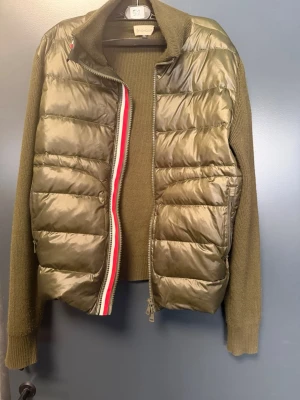 Moncler cardigan  - Snygg olivgrön cardigan från Moncler med quiltad front och stickade ärmar samt rygg. Dragkedja med röd, vit och blå detalj. Perfekt för lager-på-lager och ger en elegant men ändå clean look. Materialmix av dun och ull för både värme och stil.