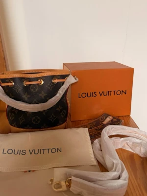  Monogram bucketbag - Snygg handväska i klassiskt monogrammönster. Väskan är i mörkbrunt och beige läder med guldfärgade metalldetaljer och dragskostängning. Kommer med originalförpackning och dustbag.