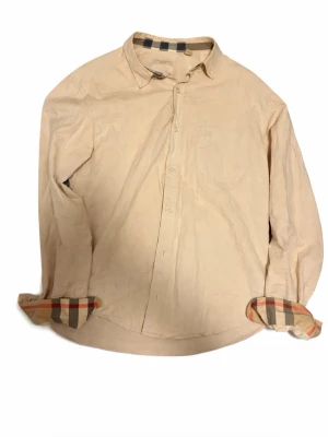 Beige skjorta från Burberry - Klassisk beige skjorta från Burberry med långa ärmar och ikoniskt rutigt mönster på insidan av krage och manschetter. Skjortan har knappar framtill och en diskret broderad logga på bröstet. Perfekt för en stilren och tidlös look.