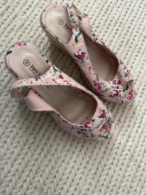 Blommiga kilklackssandaler från bpc - Säljer ett par söta sandaler från bpc med kilklack i rep och öppen tå. Skorna är ljusrosa med blommigt mönster i rosa, grönt och vitt. Justerbar rem med spänne runt hälen. Perfekta för sommarens outfits! Oanvända . 