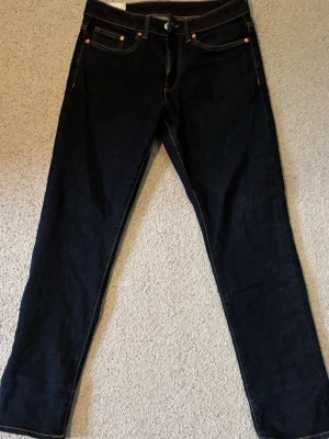 Mörkblå raka jeans med kontrastsömmar - Säljer ett par mörkblå jeans med raka ben och klassisk femficksdesign. Jeansen har kontrastsömmar och metallnitar vid fickorna. Materialet är kraftig denim som ger en tidlös look. Perfekta till en avslappnad stil och passar till det mesta.