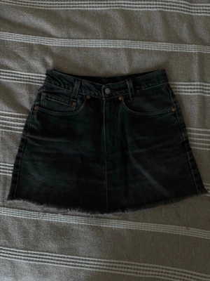 Svart jeanskjol från Levi's - Säljer en svart jeanskjol från Levi's med rå kant nertill och klassiska fem fickor. Kjolen har bälteshällor, knapp och dragkedja framtill samt Levi's-lapp bak i midjan. Perfekt för dig som gillar en edgy och avslappnad stil.