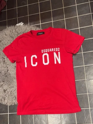 Röd ICON t-shirt från Dsquared2 - Snygg röd t-shirt från Dsquared2 med stor vit 'ICON'-text framtill. Klassisk passform och rund halsringning. Tillverkad i 100% bomull för en mjuk och bekväm känsla. Perfekt för dig som vill sticka ut med en trendig och ikonisk look.
