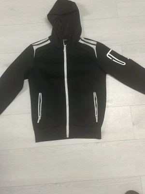 Svart hoodie med vita detaljer - Snygg svart hoodie med dragkedja framtill och vita detaljer längs dragkedjan, fickorna och axlarna. Har två fickor med dragkedja och en vit detalj på ena ärmen. Tillverkad i mjukt material och har en klassisk huva.