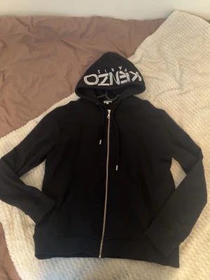 Svart Kenzo hoodie med dragkedja - Svart hoodie från Kenzo med vit logga 'KENZO PARIS' tryckt på huvan. Tröjan har dragkedja framtill, dragsko i huvan och ribbade muddar vid ärmslut och nederkant. Perfekt för en stilren och trendig look. 
