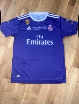 Real Madrid matchtröja lila Adidas - Säljer en lila Real Madrid matchtröja från Adidas med vita detaljer och tryck. Tröjan har texten 'FINAL CARDIFF 2017' och 'Fly Emirates' på bröstet samt klubbmärke och Adidas-logga. Korta ärmar och rund halsringning. Perfekt för fotbollsfans!