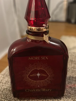 Charlotte Tilbury More Sex parfym - Lyxig parfymflaska i mörkrött glas från Charlotte Tilbury med gulddetaljer och spetsig kork. Flaskan har ett elegant guldmönster med läppar och texten 'More Sex'. Perfekt för dig som vill ha något unikt och snyggt på parfymhyllan.