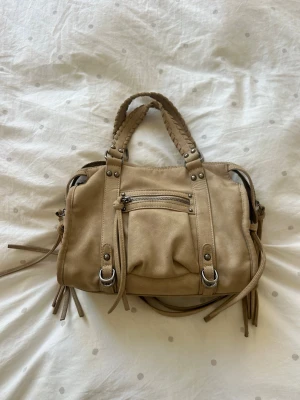 Beige handväska med flätade handtag💖💖💖 - Snygg beige handväska i mockaimitation med flätade handtag och silverfärgade metalldetaljer. Väskan har en framficka med dragkedja, dekorativa remmar och tofsar på sidorna. Perfekt för dig som gillar en trendig och stilren look.