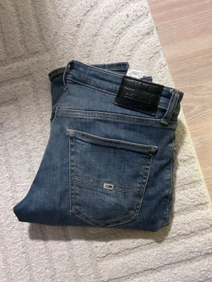 Blå Tommy Hilfiger slim jeans - Snygga blå slim fit jeans från Tommy Hilfiger med klassisk femficksdesign och diskreta slitningar. Jeansen har en svart läderpatch med logga bak i midjan och röda detaljer vid fickorna. Perfekta för en avslappnad och trendig look.