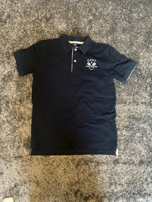 Marinblå pikétröja från Gant - Snygg marinblå pikétröja från Gant med klassisk krage och två knappar framtill. På bröstet finns ett vitt tryck med text och tennismotiv. Perfekt till en sportig och stilren look.