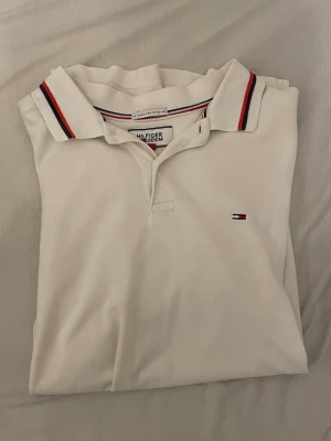 Vit pikétröja från Tommy Hilfiger - Vit pikétröja från Tommy Hilfiger med klassisk krage och röda samt marinblå detaljer på kragen. Liten broderad logga på bröstet och gråa muddar vid ärmsluten. Perfekt för en stilren och sportig look.