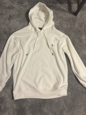 Vit hoodie från Polo Ralph Lauren - Snygg vit hoodie från Polo Ralph Lauren med klassisk känguruficka och dragsko i huvan. Liten broderad logga på bröstet. Mjuk bomullskänsla och stilren design som passar perfekt till jeans eller joggers.