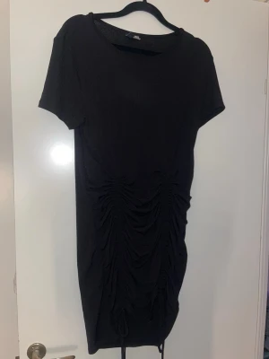 Svart kortklänning med snörning - Svart kortklänning från H&M med korta ärmar och snygga snörningsdetaljer framtill som ger en rynkad effekt. Klänningen har en enkel rund halsringning och är tillverkad i ett mjukt material som känns bekvämt mot huden.