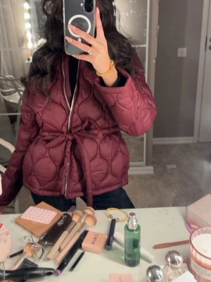 Vinröd pufferjacka med knytband - Snygg vinröd pufferjacka med quiltat mönster och knytband i midjan. Jackan har rund halsringning, dragkedja framtill och långa ärmar. 