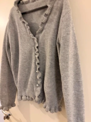 Grå volangstickad kofta - Supersöt grå kofta med volangkant längs knäppningen, ärmslut och nederkant. Koftan har långa ärmar och pärlknappar framtill. 