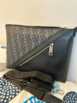 Svart axelväska från Fendi - Säljer en svart axelväska från Fendi med ikoniskt FF-mönster i grått och svart. Väskan har en diagonal dragkedja framtill och justerbar axelrem med Fendi-logga. Materialet är en mix av läder och canvas, perfekt för dig som gillar lyxiga detaljer och streetstyle.