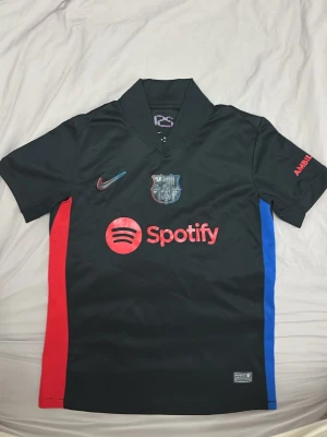 FC Barcelona svart matchtröja Nike - Snygg svart FC Barcelona matchtröja från Nike med klubbmärke, Spotify-logga och röda samt blå detaljer på sidorna. Kortärmad med krage och tryck på både fram- och baksida. Perfekt för dig som älskar fotboll och vill representera Barca. OBV! Nike loggan är lite lös.