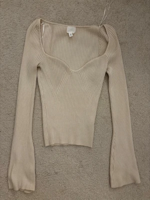 Beige ribbad långärmad topp - Trendig beige ribbad topp med lång ärm och markerad byst. Toppen har en fin passform och bred, fyrkantig urringning som ger en modern look. Perfekt att styla med höga jeans eller kjol.