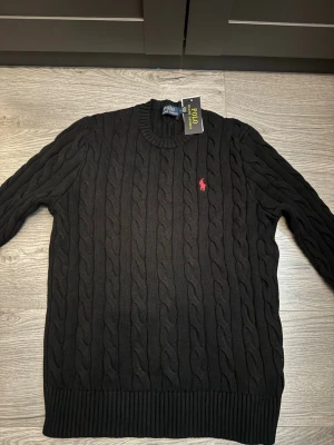 Svart kabelstickad tröja Polo Ralph Lauren - Kabelstickad svart tröja från Polo Ralph Lauren med rund halsringning och röd broderad logga på bröstet. Tröjan har ribbade muddar vid ärmslut och nederkant. Klassisk och stilren design som passar till mycket.