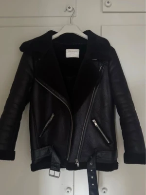 Svart skinnjacka med bälte Zara - Snygg svart skinnjacka från Zara med bred krage, dragkedjor i silver och bälte i midjan. Jackan har två snedställda fickor med dragkedja och en cool bikerstil. Perfekt för dig som vill ha en trendig och tidlös look.