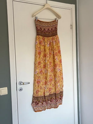 Gul blommig långklänning med smock - Superfin långklänning i gul bas med rosa och orange blommigt mönster. Klänningen har smock upptill och breda bårder med broderade detaljer i överdelen och nederkanten. Köpte den själv på Vinted förra sommaren så vet tyvärr inte var den är från