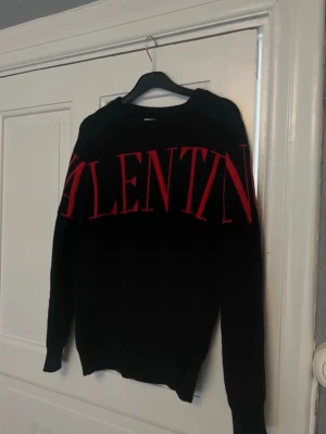 Svart stickad tröja från Valentino - Snygg svart stickad tröja från Valentino med stort rött logotyp-tryck över bröstet. Tröjan har rund halsringning och långa ärmar. Perfekt för dig som gillar statement-plagg och exklusiv design.