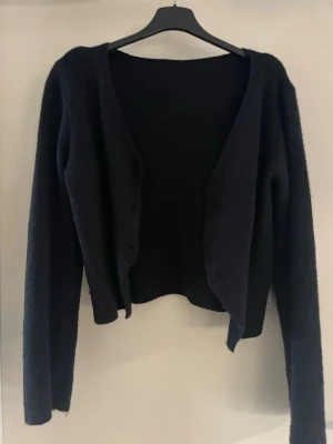 Svart croppad kofta med lång ärm - Säljer en svart, croppad kofta med lång ärm och enkel knäppning framtill. Koftan har en mjuk och lite fluffig yta, perfekt att slänga över en topp. Enkel och stilren design som passar till det mesta.