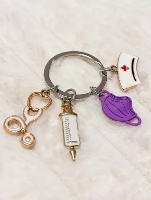 Nyckelring med vårdtema och berlocker - Nyckelring i metall med fyra berlocker: stetoskop i guld, spruta i vitt och guld, lila munskydd och vit sjuksköterskehatt med röd detalj. Perfekt accessoar för dig som gillar vård eller pluggar till sjuksköterska.