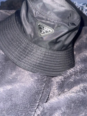 Svart bucket hat från Prada - Snygg svart bucket hat från Prada i glansigt nylonmaterial med klassisk trekantig logotyp framtill. Perfekt accessoar för att lyfta din streetwear-look och ge outfiten en exklusiv touch.