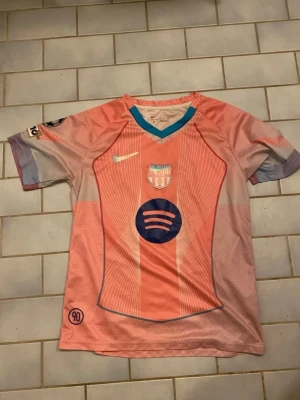 Rosa FC Barcelona matchtröja Nike - Säljer en rosa och ljusblå FC Barcelona matchtröja från Nike med klubbmärke och Spotify-logga på bröstet. Tröjan har korta ärmar, V-ringning med blå kant och detaljer i vitt och lila. Perfekt för dig som älskar fotboll och vill sticka ut på planen.